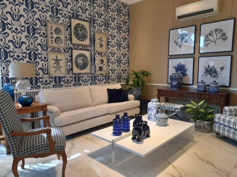 Sala sofisticada com parede azul estampada, quadros decorativos com conchas e estrelas-do-mar, porcelanas azuis e brancas em destaque na decoração.