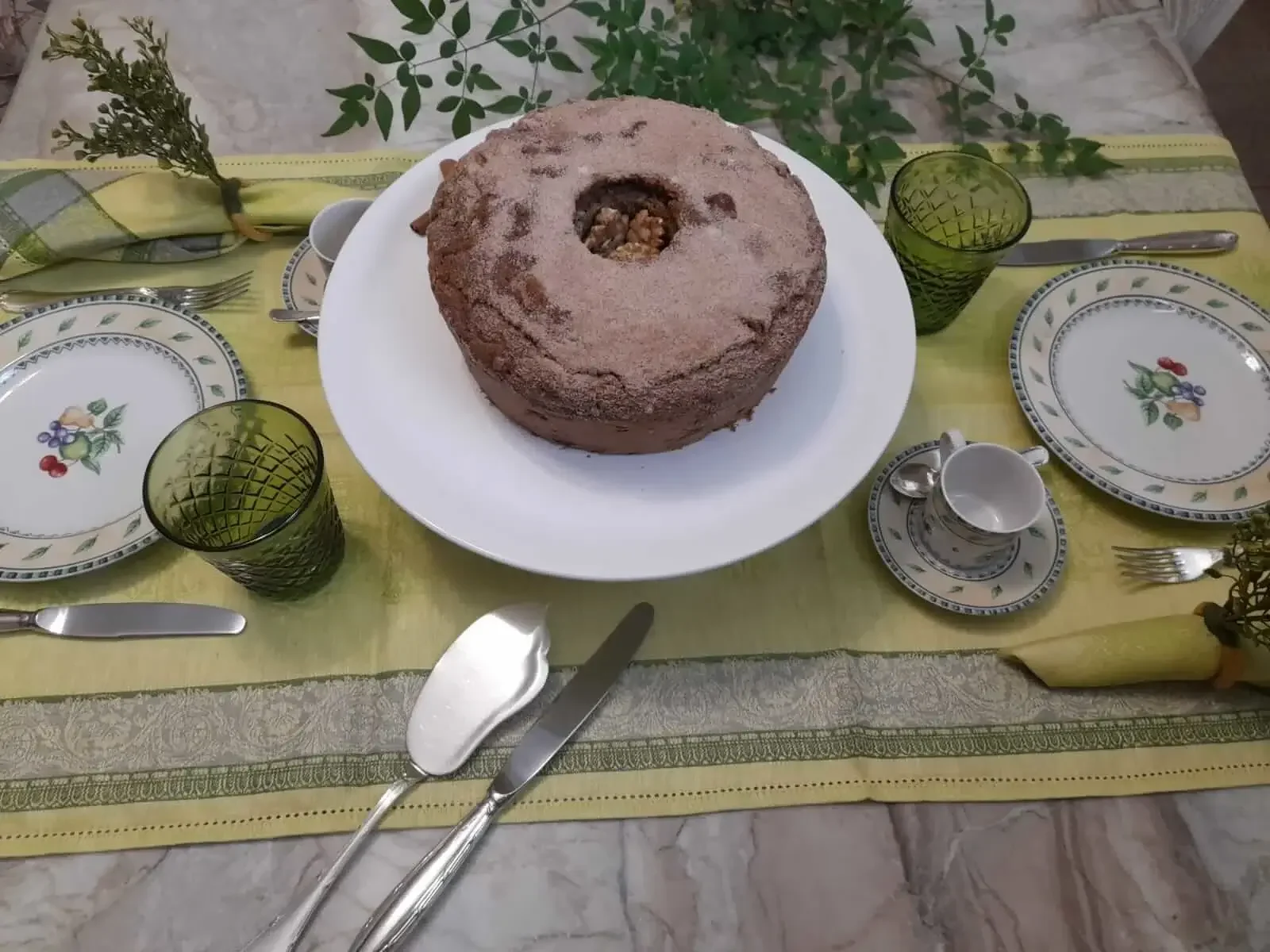 Mesa com trilho português verde, bolo de maçã e canela sobre boleira branca e louças delicadas para café da tarde elegante.