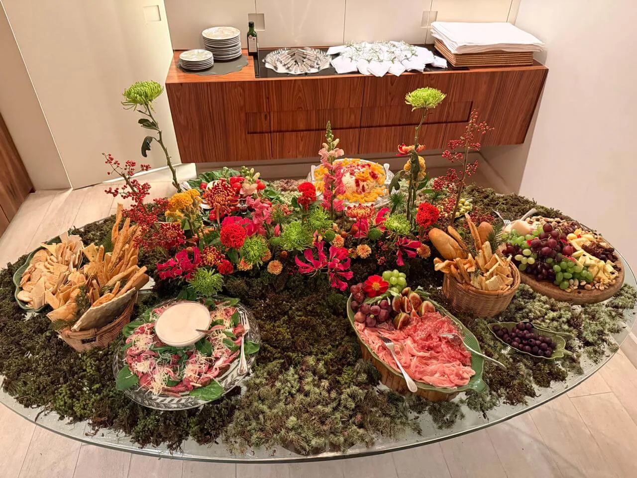 Mesa decorada para receber amigos com flores coloridas, musgo natural, queijos, frutas, pães artesanais e antepastos inspirada em um jardim.